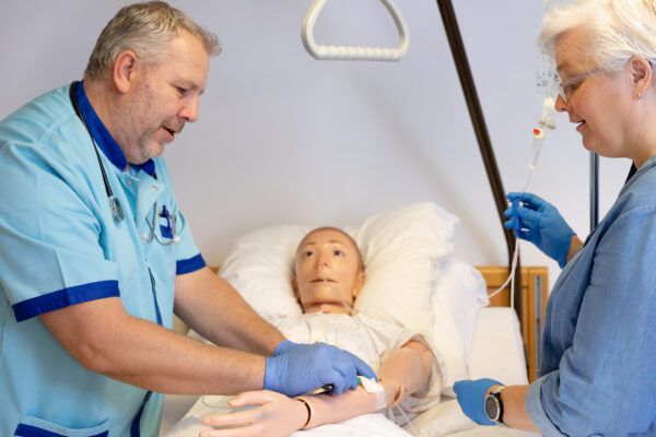 Docent en student volwassenenonderwijs zorg oefenen samen met simulatiepatient in ziekenhuisbed