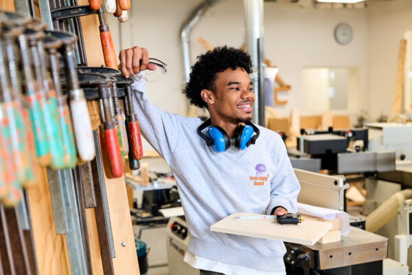 Mbo-student Bouw en Techniek staat voor een wand met gereedschap en kijkt blij.