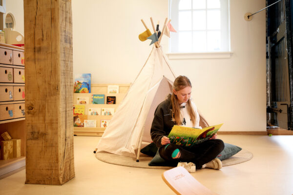 Mbo-student pedagogisch medewerker kinderopvang zit op de grond in een speelgoedtentje en leest een boek